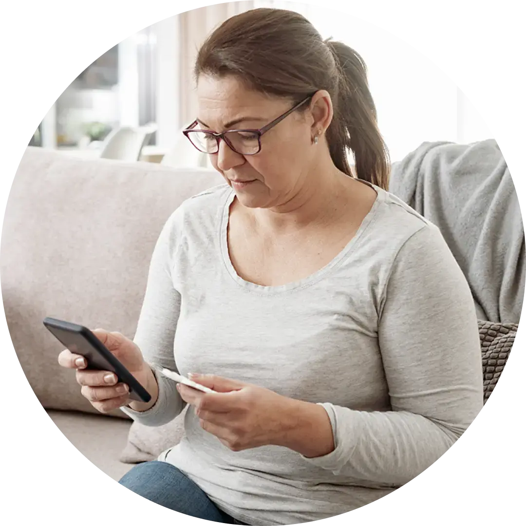 A woman with glasses sitting on a sofa, looking at her smartphone while holding a small white card. She is in a bright, neutral-toned living room, and the image is cropped into a circular frame.