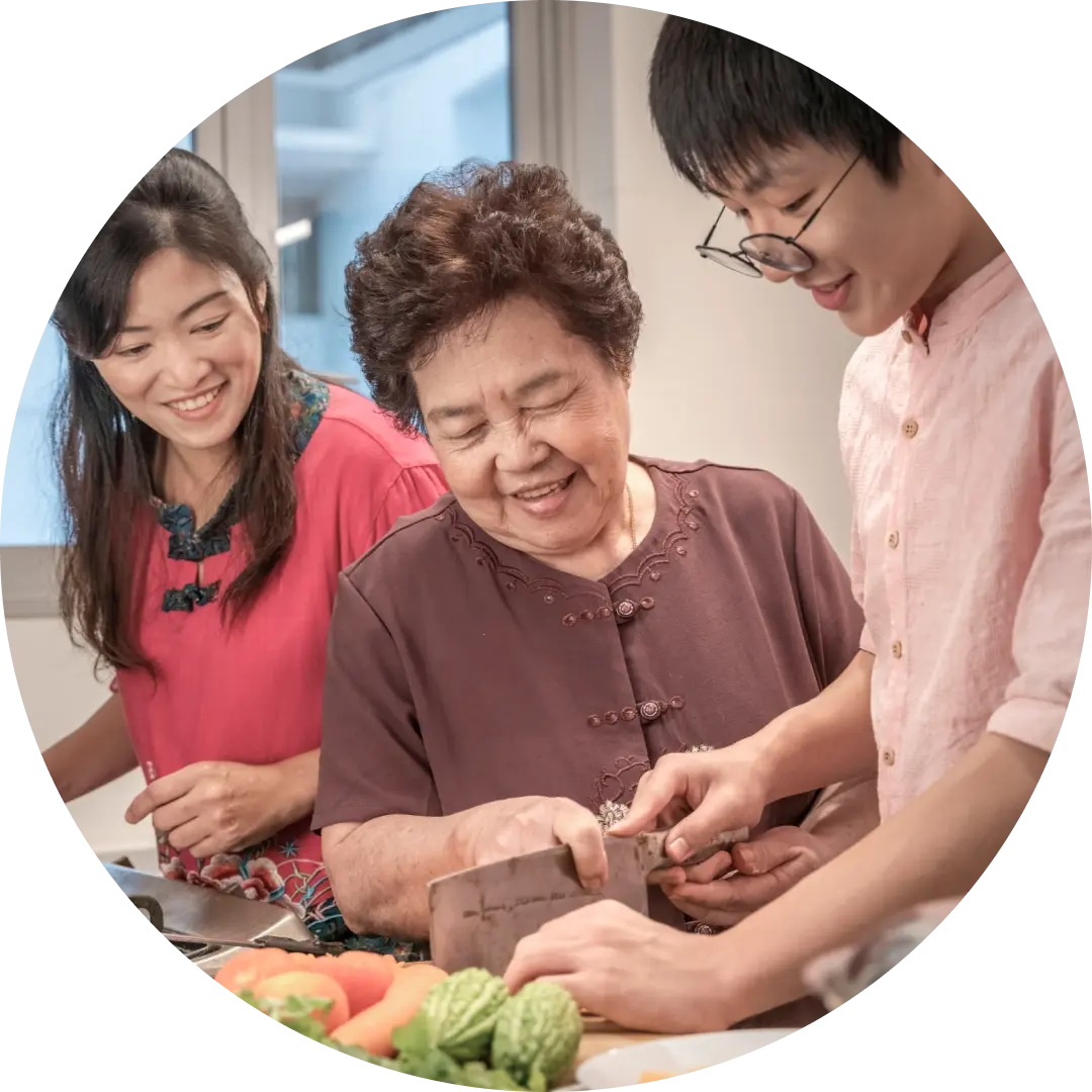A multi-generational Asian family prepares food together in a kitchen, smiling and cutting vegetables with a cleaver..